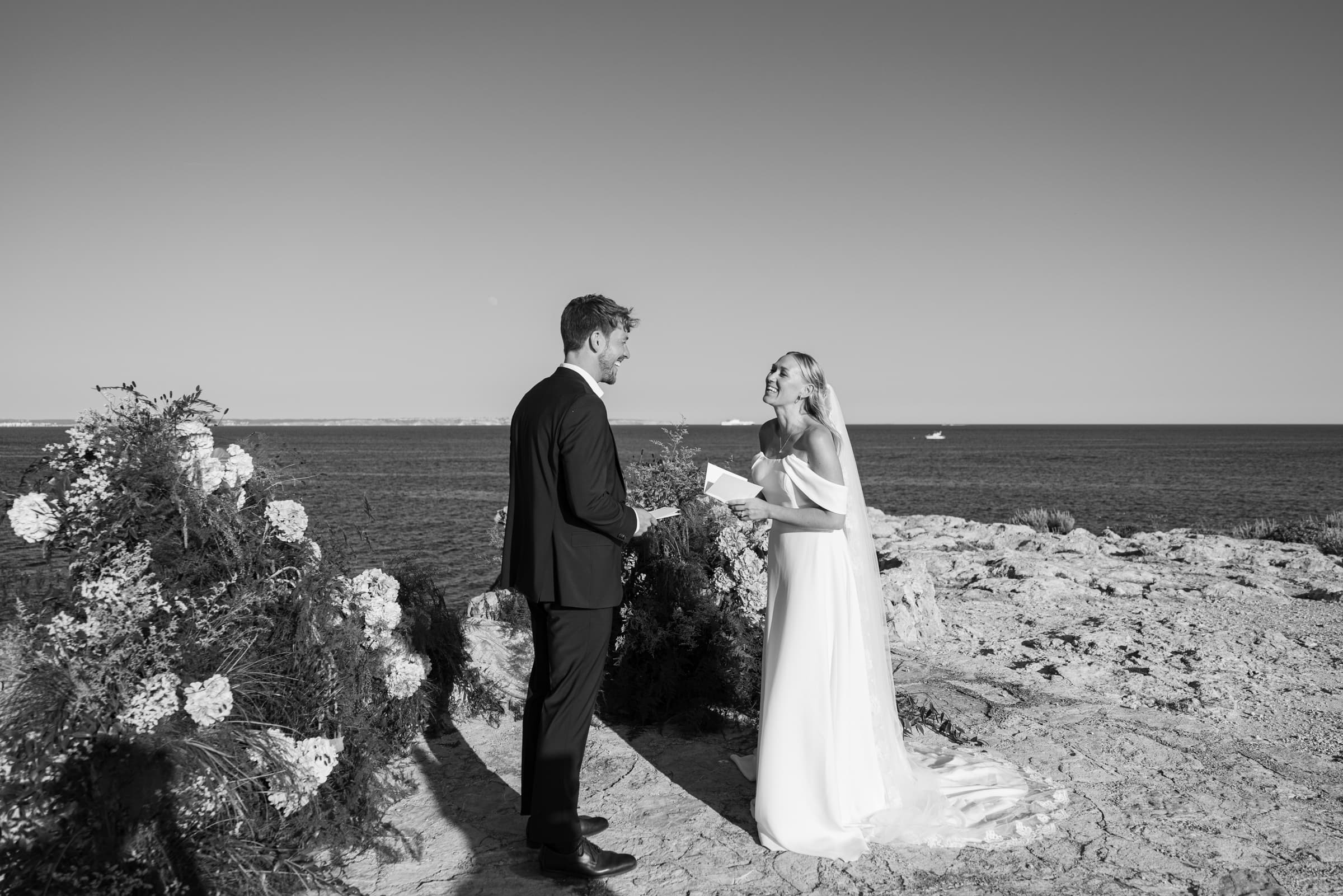 Black-and-white wide view of a couple in wedding attire beside flowers on a rocky coast.
