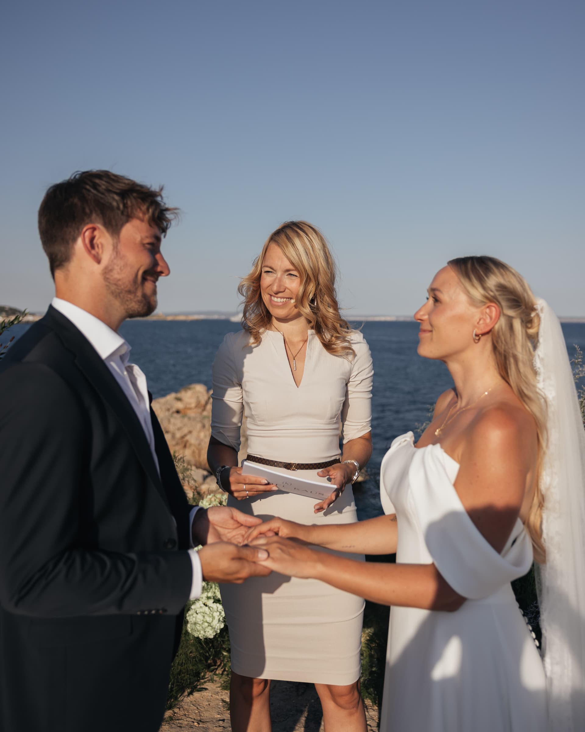 A couple in wedding attire holding hands with a woman beside the sea.