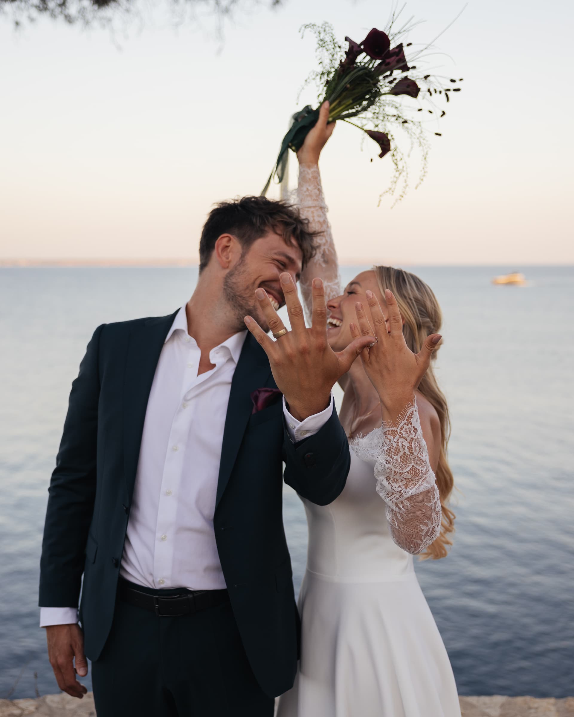 Ein frisch verheiratetes Paar lacht am Meer, während die Person in Weiß einen Blumenstrauß hebt.