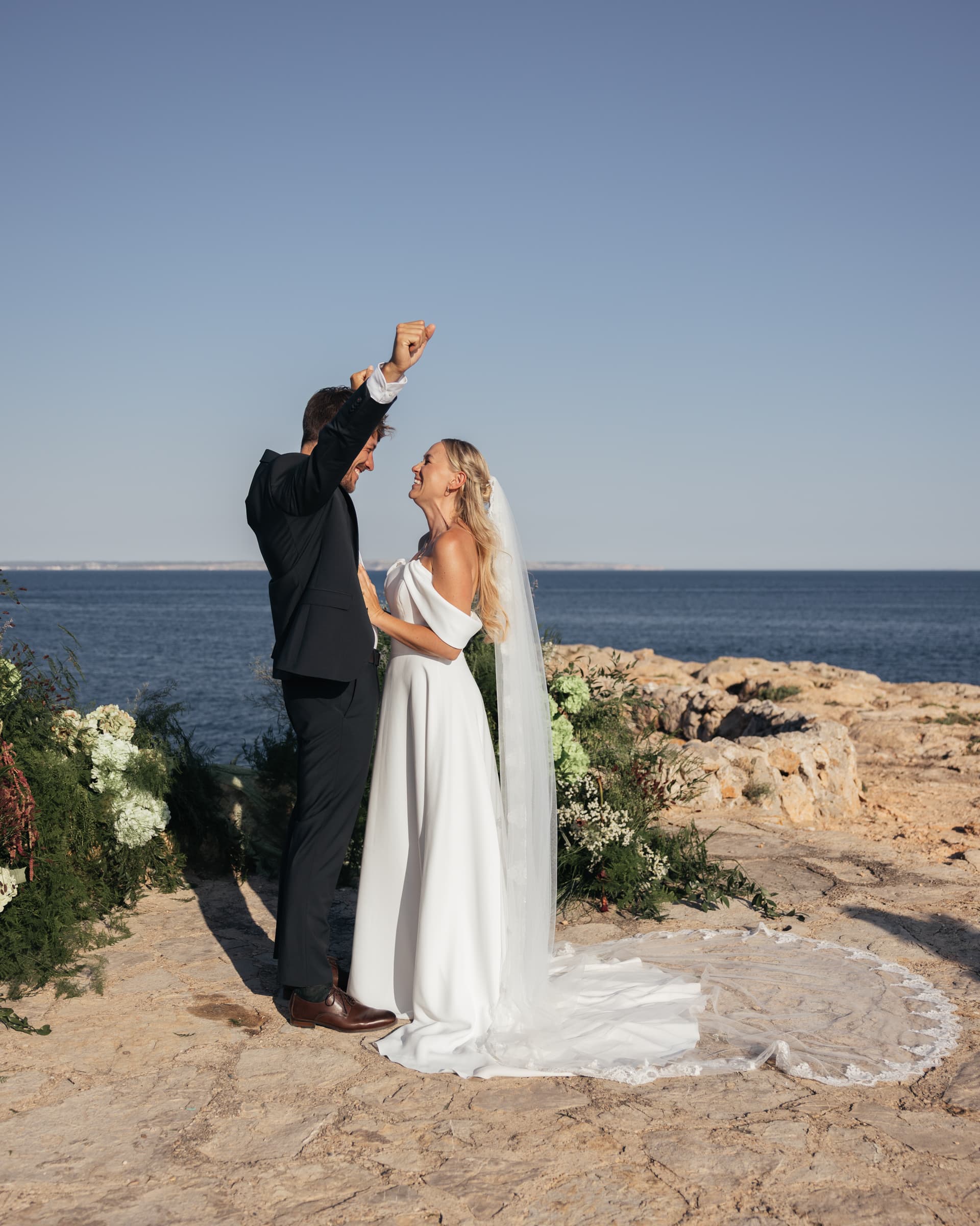 A couple in wedding attire standing beside flowers on a rocky coast.
