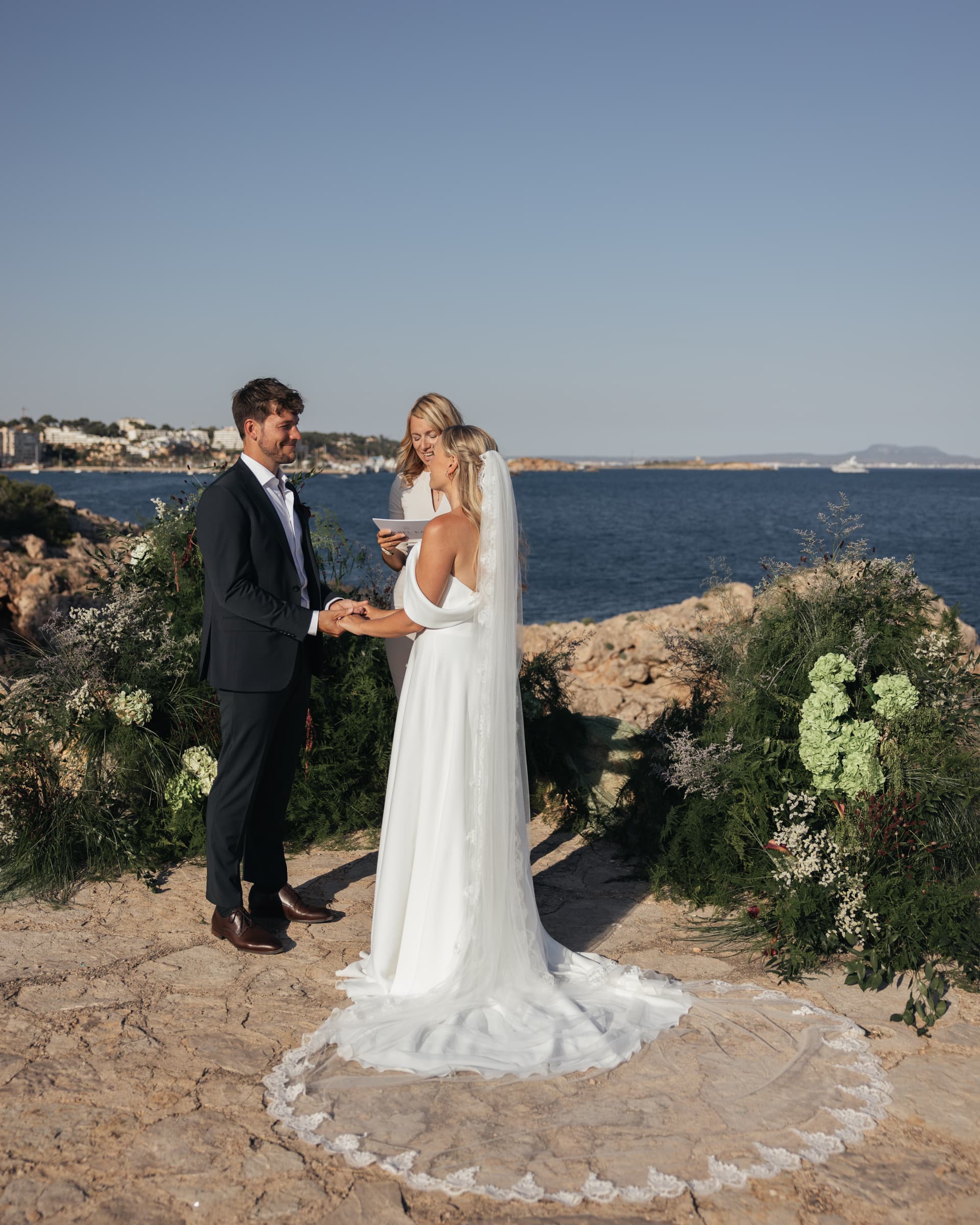 Wide view of a couple in wedding attire outdoors with the sea behind.