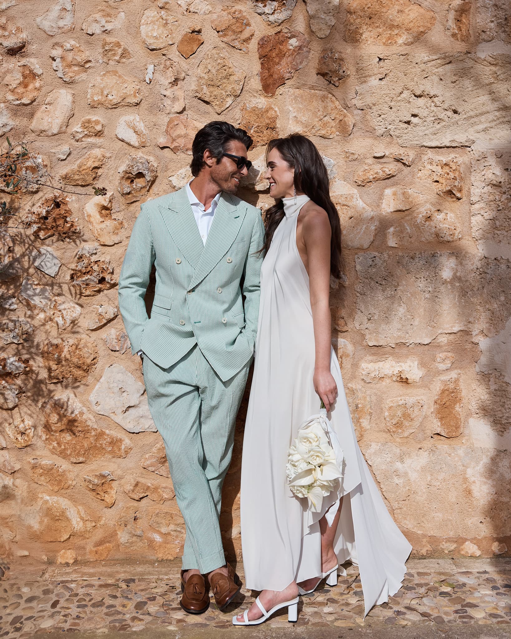 A couple in wedding attire standing beside a textured stone wall.