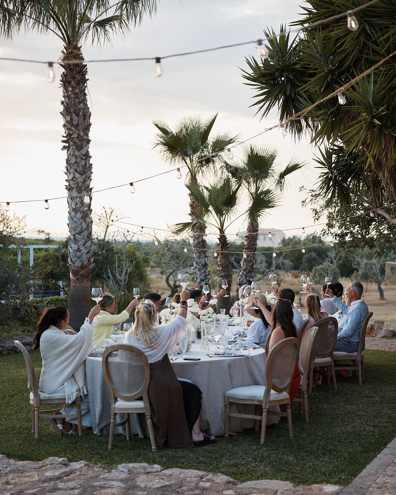 A long outdoor dinner table beneath palm trees and string lights.