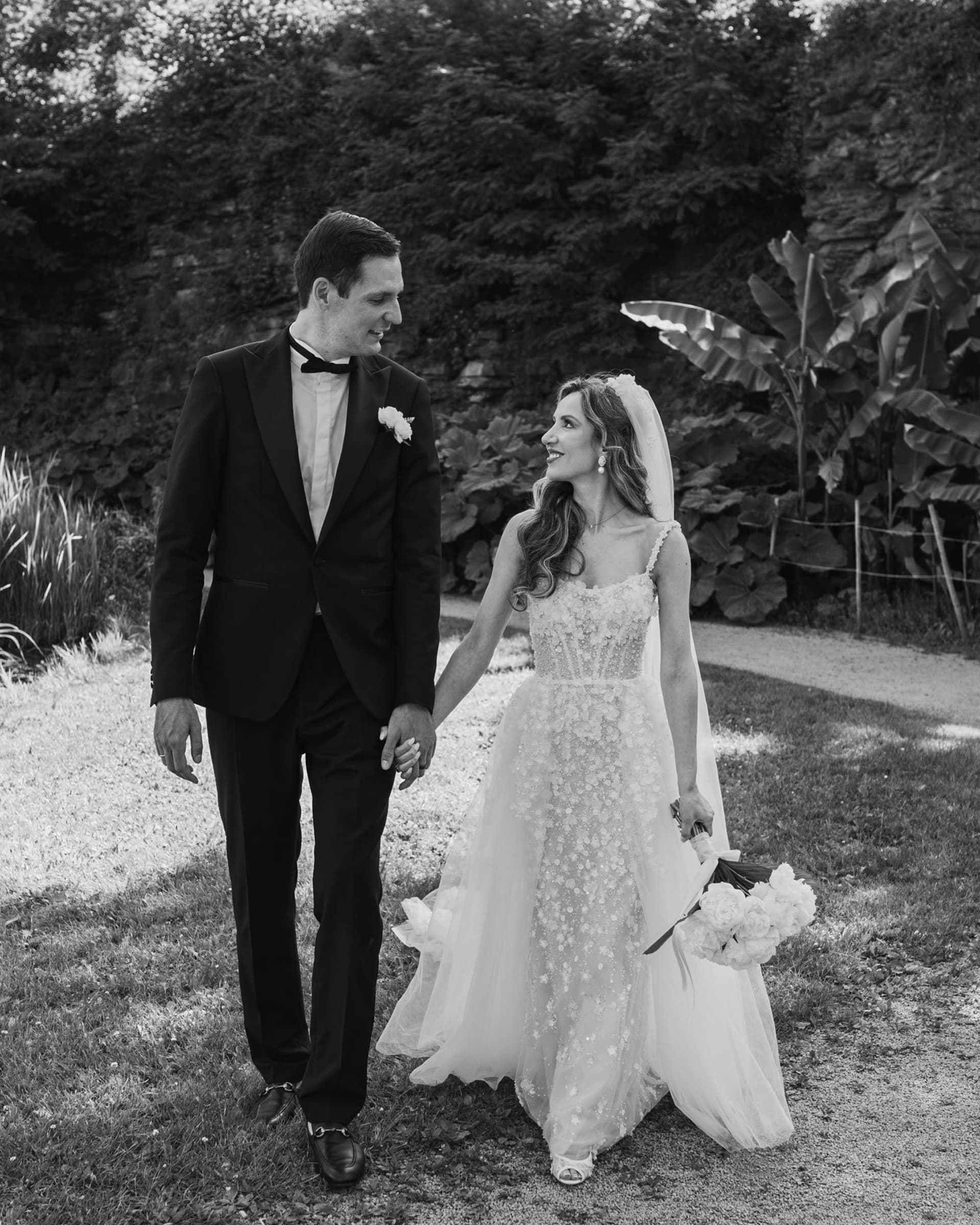 Black-and-white photo of a couple in light formal clothing walking together on a rocky shoreline with the sea behind them.
