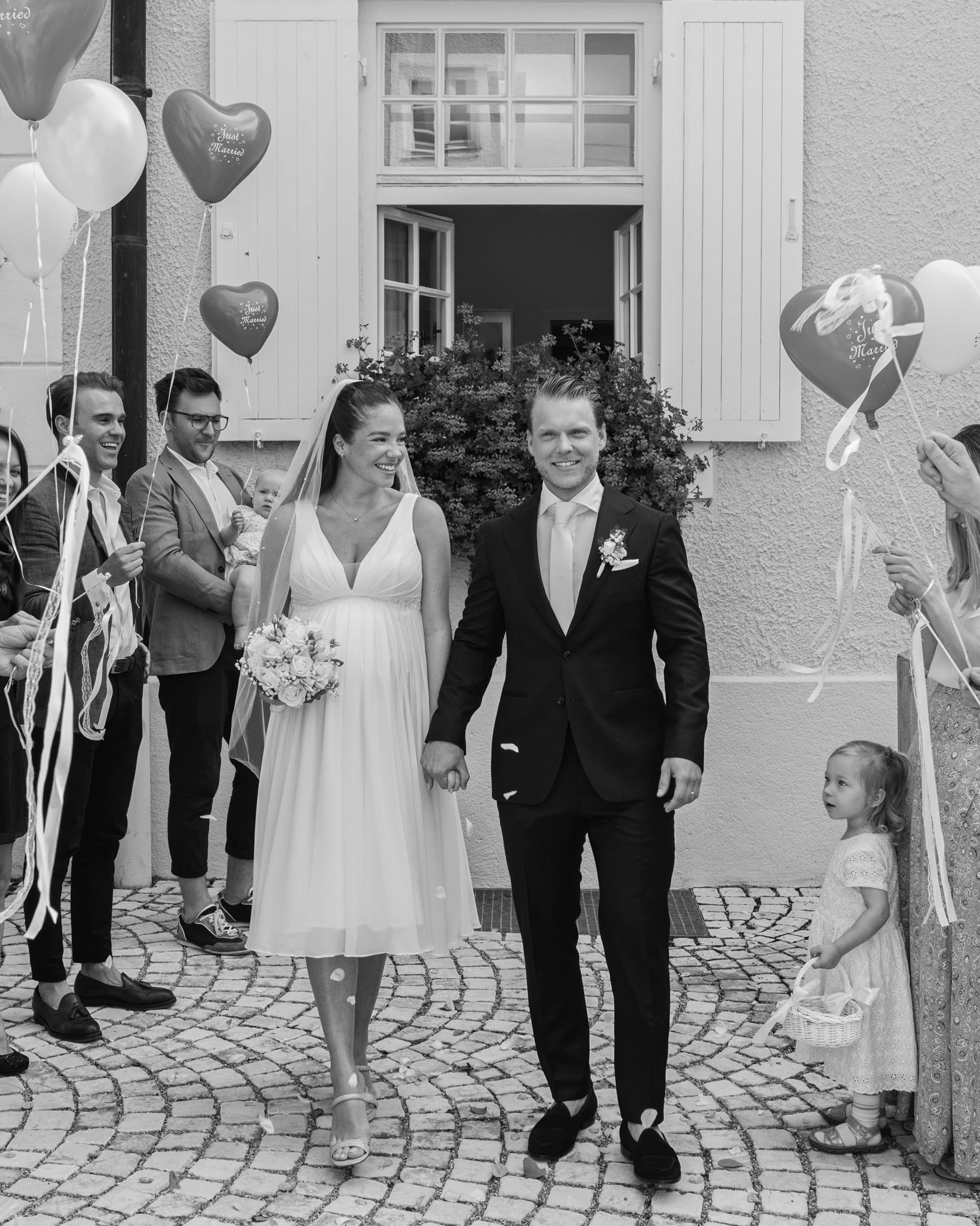 Black-and-white photo of a couple in wedding attire walking outdoors with people behind them.